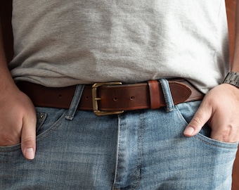 Puede incluir: Un cinturón de cuero marrón con una hebilla de latón. El cinturón se lleva alrededor de la cintura de una persona que lleva pantalones vaqueros azules.
