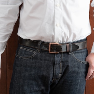 May include: A brown leather belt with a copper buckle. The belt is worn around the waist of a person wearing a white button-down shirt and dark blue jeans.