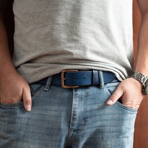 May include: A person wearing blue jeans with a dark blue leather belt with a copper buckle. The person is wearing a grey t-shirt and a silver watch.