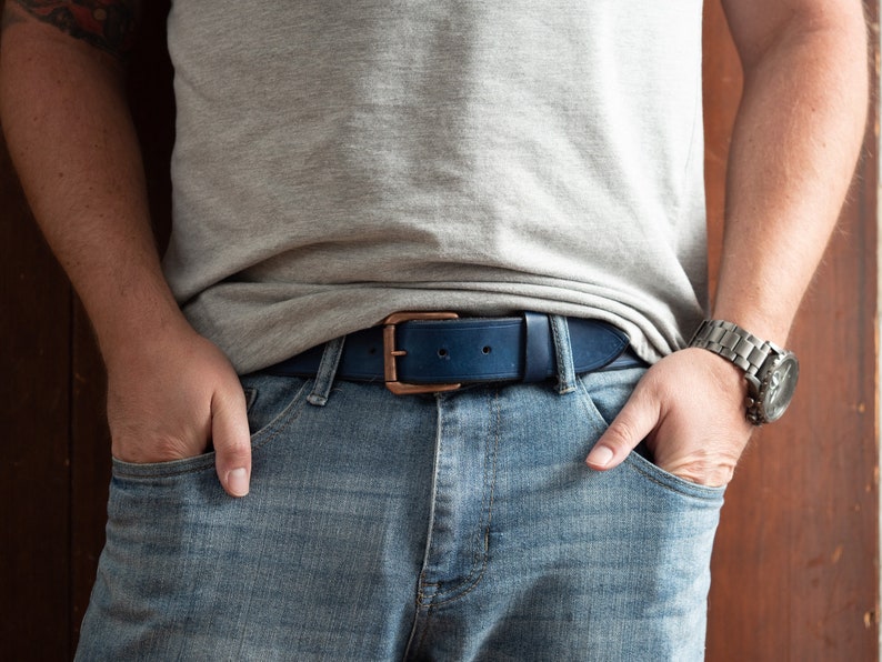 May include: A person wearing blue jeans with a dark blue leather belt with a copper buckle. The person is wearing a grey t-shirt and a silver watch.