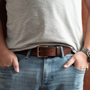 May include: A brown leather belt with a copper buckle. The belt is worn over blue jeans.