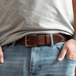 May include: A brown leather belt with a copper buckle. The belt is worn around the waist of a person wearing blue jeans.