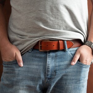 May include: A brown leather belt with a silver buckle. The belt is worn around the waist of a person wearing blue jeans and a grey t-shirt.