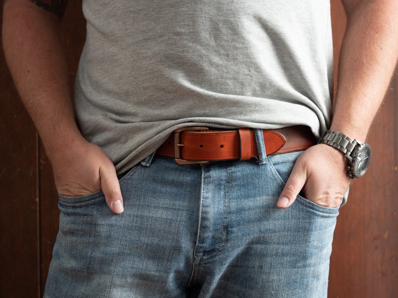 May include: A brown leather belt with a silver buckle. The belt is worn around the waist of a person wearing blue jeans and a grey t-shirt.