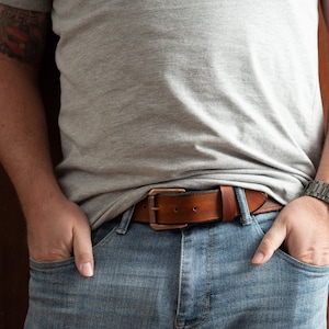 May include: A brown leather belt with a copper buckle. The belt is worn over a pair of blue jeans.