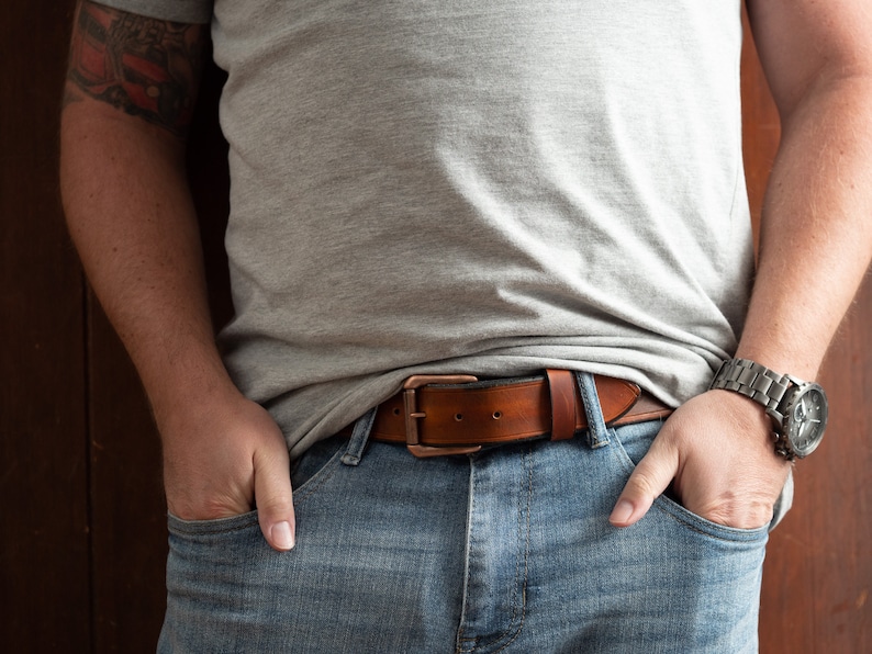May include: A brown leather belt with a copper buckle. The belt is worn over a pair of blue jeans.