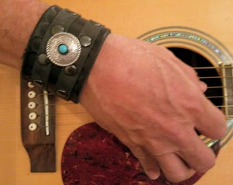 May include: A black leather cuff bracelet with a silver and turquoise concho. The bracelet is worn on a person's wrist.