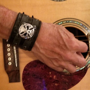 Puede incluir: Una pulsera de cuero negro con un diseño de calavera y huesos cruzados plateados. La pulsera se lleva en la muñeca de una persona, y la persona está tocando una guitarra acústica.