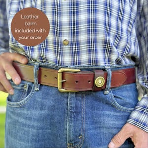 May include: A brown leather belt with a brass buckle is fastened on a person wearing blue jeans and a plaid shirt. The belt has a loop with a decorative brass button. The text "Leather balm included with your order" is in a brown circle.