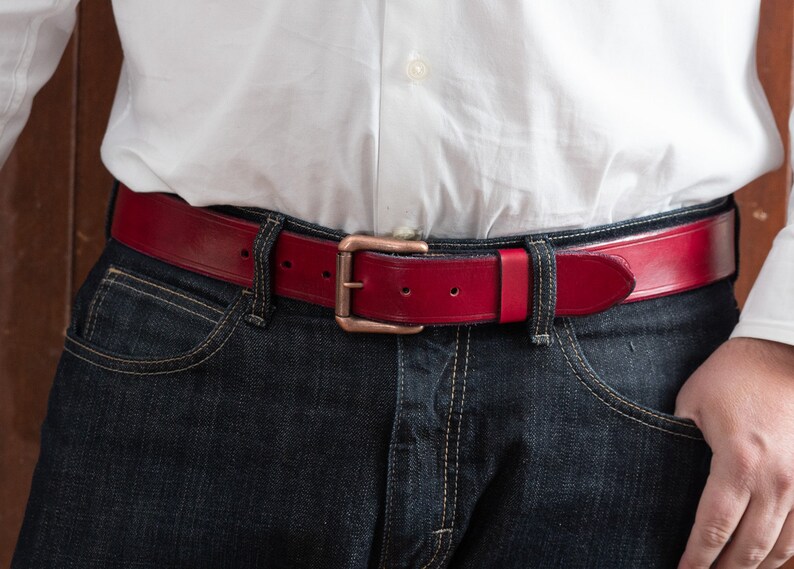 May include: A red leather belt with a copper buckle. The belt is worn over a pair of dark blue jeans.