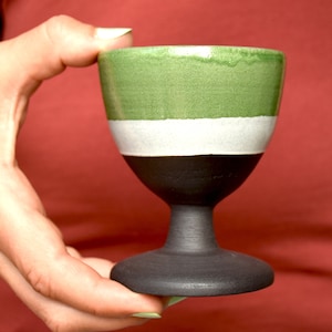 Könnte beinhalten: Ein handgefertigter Keramikbecher mit grünem Oberteil, weißem Streifen und schwarzem Boden. Der Becher wird von einer Person gehalten. Der Becher hat einen breiten Boden und einen kurzen Stiel. Der Hintergrund ist einfarbig rot.