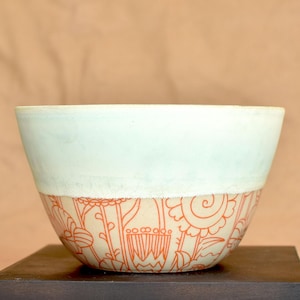 Puede incluir: Un cuenco de cerámica con una sección superior azul claro y una sección inferior decorada con patrones florales naranjas. El cuenco está sobre una superficie marrón oscuro sobre un fondo neutro.