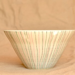 Puede incluir: Un cuenco de cerámica de color crema con forma acampanada. El cuenco está decorado con rayas verticales en tonos rosa y verde. El cuenco se utiliza probablemente para servir alimentos.