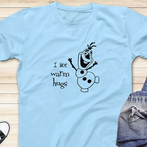 Puede incluir: Camiseta azul claro con un mu&ntilde;eco de nieve de dibujos animados negro y el texto "Me gustan los abrazos c&aacute;lidos".
