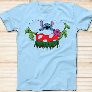 May include: Light blue t-shirt featuring a cartoon character design. The design includes a blue alien character, a red and white mushroom, green leaves, and a pink flower. The shirt is a crew neck style.