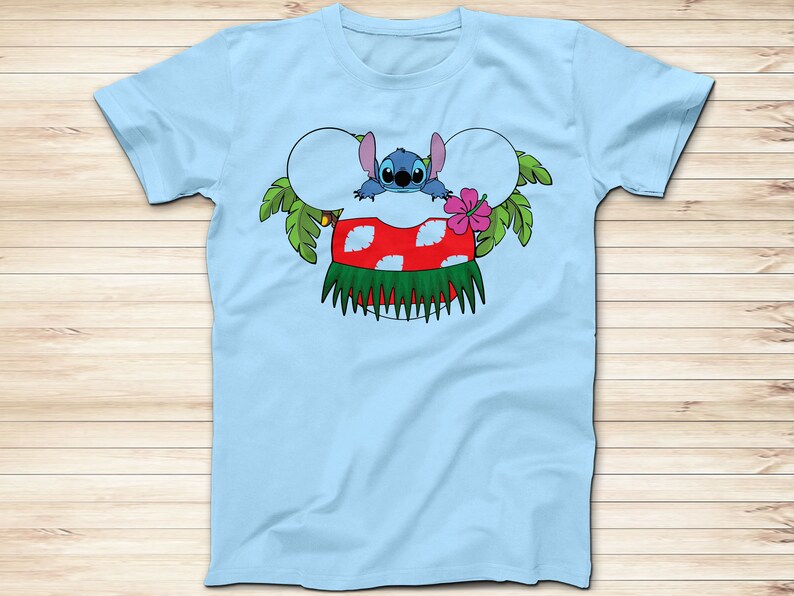 May include: Light blue t-shirt featuring a cartoon character design. The design includes a blue alien character, a red and white mushroom, green leaves, and a pink flower. The shirt is a crew neck style.
