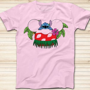 May include: A light pink t-shirt featuring a cartoon design of Stitch from Lilo & Stitch. Stitch is depicted with mouse ears, a red and white mushroom, and a green grass skirt. A pink hibiscus flower and green leaves complete the design.