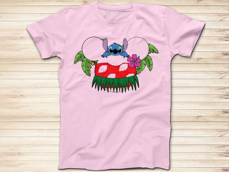 May include: A light pink t-shirt featuring a cartoon design of Stitch from Lilo & Stitch. Stitch is depicted with mouse ears, a red and white mushroom, and a green grass skirt. A pink hibiscus flower and green leaves complete the design.