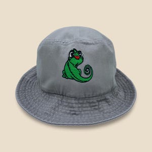 May include: A gray bucket hat featuring an embroidered green chameleon with a red tongue. The hat has a rounded crown and a wide brim. The chameleon is in a playful pose, adding a whimsical touch to the casual hat.