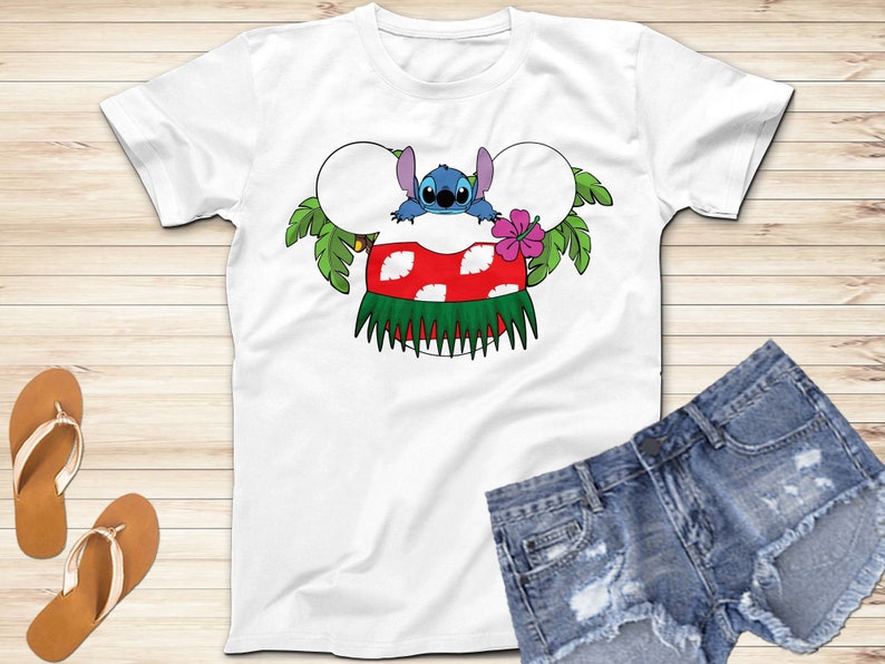 May include: White t-shirt featuring a cartoon design of Stitch from Lilo & Stitch. The design includes Stitch peeking over a red and white mushroom with a green grass skirt, surrounded by palm leaves and a pink flower. Denim shorts and sandals are also visible.