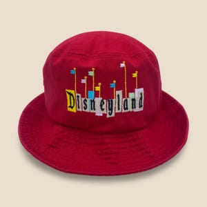 May include: A red bucket hat with the word "Disneyland" embroidered in colorful letters and flags. The hat is made of a soft fabric and has a wide brim. A classic souvenir.