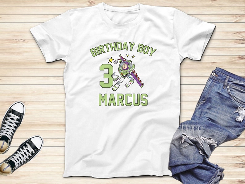 May include: White t-shirt with a green and purple graphic of a cartoon character holding a toy rocket. The text "Birthday Boy" is above the character and the number "3" is to the left of the character. The name "Marcus" is below the character.
