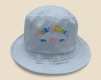 May include: A light blue bucket hat with the text "Disney World" embroidered in pink, yellow, and teal. The hat also features a pink Mickey Mouse head and the text "EST. 1971". The hat has a wide brim and is made of a soft fabric.