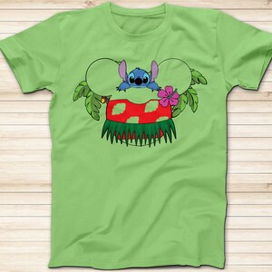May include: A light green t-shirt featuring a cartoon graphic of Stitch from Lilo & Stitch. Stitch is depicted with a Hawaiian lei and a red and green hula skirt. The design includes Mickey Mouse ears and tropical leaves.