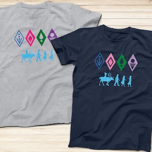 Op de afbeelding: Twee T-shirts met een grafische print van een rendier en vier figuren. De T-shirts zijn grijs en donkerblauw. De grafiek bevat vijf ruitvormige symbolen in blauw, roze, groen en paars. De tekst op de T-shirts luidt "Let It Go".