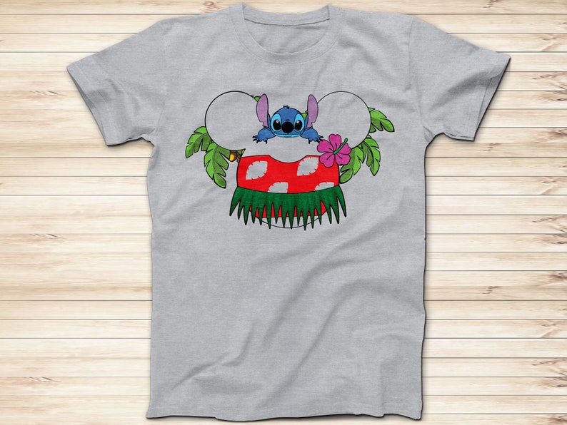 May include: A light gray t-shirt featuring a colorful graphic of the character Stitch from Lilo & Stitch. Stitch is depicted in a Hawaiian-themed design, with a red and white mushroom, green grass skirt, and a pink flower. The design is in the shape of Mickey Mouse ears.