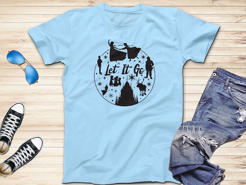 Op de afbeelding: Lichtblauw T-shirt met een zwart silhouetontwerp van personages uit de film Frozen. Het ontwerp bevat de woorden "Let It Go" en een sneeuwvlok.