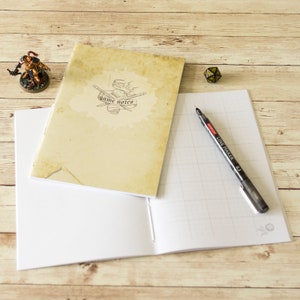May include: A notebook with a vintage-style cover featuring the text "game notes" and a drawing of dice and pencils. The notebook is open to a blank page with a grid pattern. A black pen is lying on the open page.