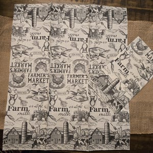 May include: A set of two farm-themed cloth napkins. The napkins feature a repeating pattern of farm scenes, including tractors, barns, cows, and the text "Farm Milk" and "Farmer's Market". The design is in black and white.