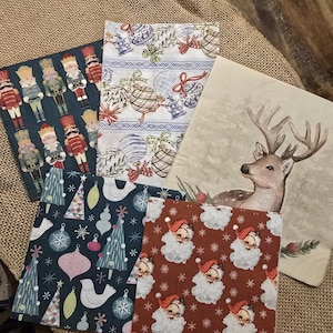 May include: A collection of decorative Christmas napkins with various festive designs. The napkins feature images of nutcrackers, ornaments, Santa Claus, Christmas trees, and a deer. The napkins are in shades of red, blue, green, and white.