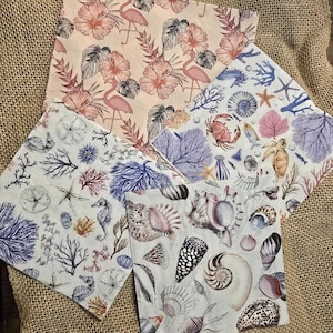 May include: Four decorative paper napkins with various coastal designs. One features pink flamingos and flowers, another has seashells, and two others display coral, starfish, and seahorses. The napkins are square and rest on a burlap surface.