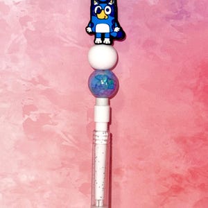 Op de afbeelding: Een pen met een blauw stripfiguur, witte en blauwe kralen en een doorzichtige barrel gevuld met glitter. De pen heeft een witte bovenkant en een zilveren basis. De achtergrond is een roze en witte gradiënt.
