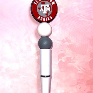 Puede incluir: Bolígrafo blanco y plateado con el logotipo de Texas A&M Aggies. El bolígrafo tiene un logotipo redondo, rojo, blanco y gris en la parte superior con las palabras "TEXAS A&M AGGIES". El bolígrafo tiene un cuerpo blanco y una punta plateada.