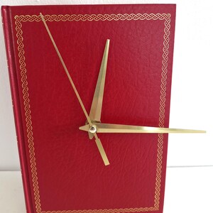 Könnte beinhalten: Eine rote, buchförmige Uhr mit goldfarbenen Zeigern und einem goldenen Rand. Die Zeiger sind dünn und spitz. Das Zifferblatt ist einfarbig rot, und der Rand hat ein sich wiederholendes Muster.