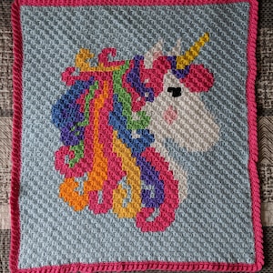 Puede incluir: Una manta de ganchillo con forma de unicornio con una melena arcoíris y un fondo azul. La manta tiene un borde rosa.