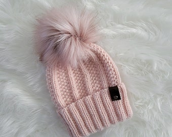 May include: A pink knitted beanie with a fluffy pom pom on top. The beanie has a ribbed texture and a small black leather tag.