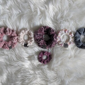 Op de afbeelding: Zes handgemaakte scrunchies in verschillende kleuren en patronen, waaronder roze, wit, paars en grijs. De scrunchies zijn gemaakt van zacht, gecrochete stof en zijn perfect om elke haarstijl een vleugje stijl te geven.