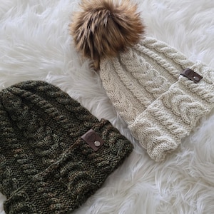 May include: Two cable-knit beanies. One is olive green with a leather tag, the other is cream-coloured with a faux fur pom-pom and a leather tag. Both beanies are on a white, fluffy surface.