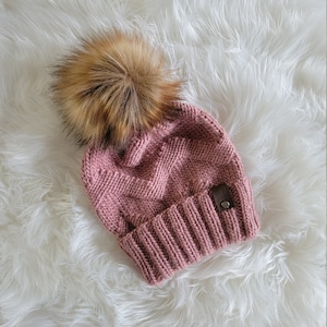 Puede incluir: Un gorro de punto rosa polvoriento con un gran pompón de piel sintética marrón y esponjoso. El gorro tiene un borde acanalado y una pequeña etiqueta de cuero marrón. El gorro se muestra sobre una superficie blanca y esponjosa.