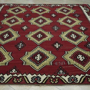 a large, intricately patterned rug with a red background and a border of white and gold designs. The rug features a repeating pattern of diamond shapes in various sizes and colors, creating a visually striking and detailed design.