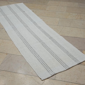 May include: A rectangular, off-white rug featuring three grey stripes along its length. The rug, made from woven material, is shown on a light-coloured tiled floor. The design of the rug is simple and minimalist.