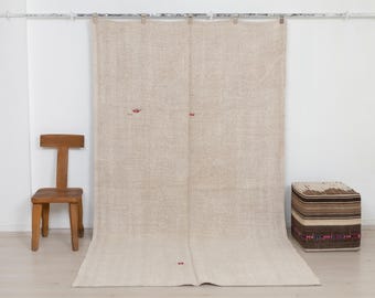 May include: A large, rectangular, beige woven textile hanging vertically, featuring a coarse, natural fiber texture with subtle, small red embroidered accents scattered across the surface, suspended from a metal rod with clips.