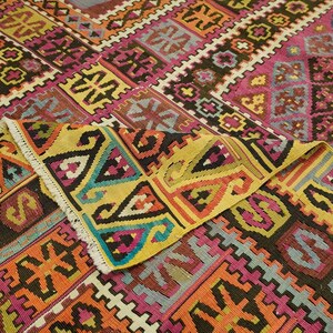 a vibrant and intricate rug with a complex geometric pattern in a variety of colors, including shades of pink, yellow, orange, and black. The rug is folded and displayed in a close-up view, showcasing the detailed design and craftsmanship.