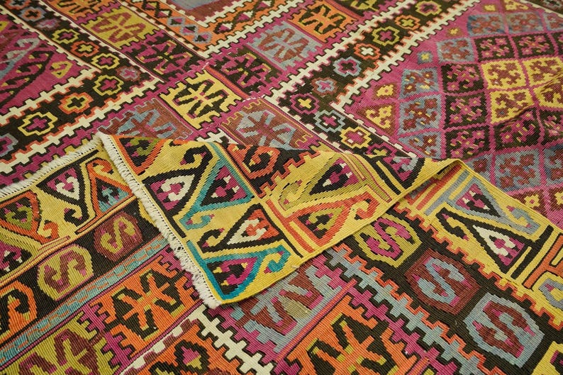 a vibrant and intricate rug with a complex geometric pattern in a variety of colors, including shades of pink, yellow, orange, and black. The rug is folded and displayed in a close-up view, showcasing the detailed design and craftsmanship.