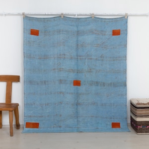 May include: A large, rectangular, light blue textile wall hanging featuring horizontal, faded brown stripes and four small, solid orange rectangular patches positioned near the corners, with one additional orange patch centered.