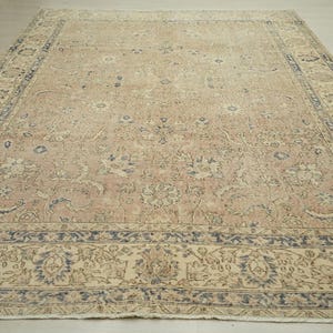 a large, intricately patterned rug with a floral design in shades of beige and blue. The rug is placed on a wooden floor, and the background is a plain white wall.